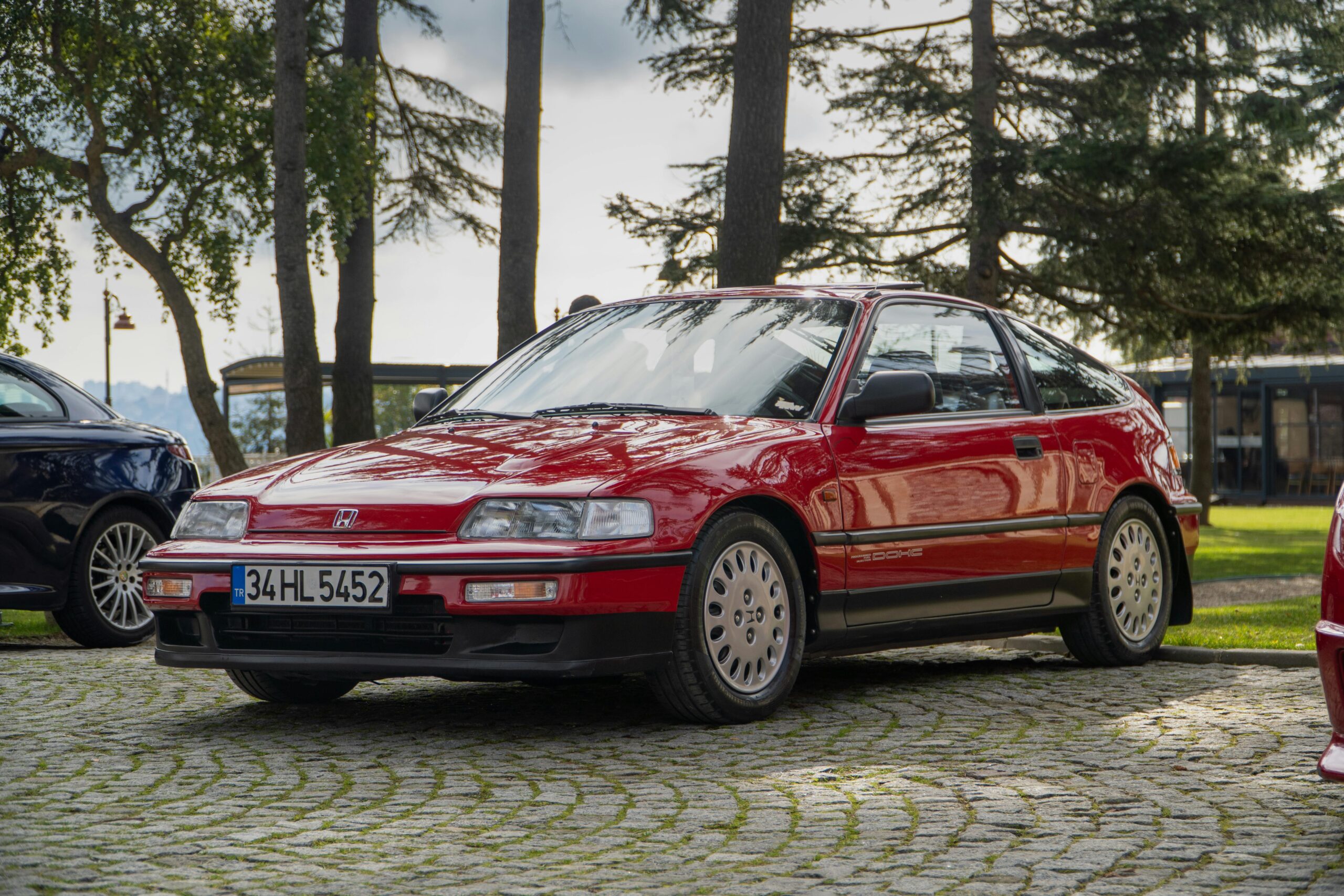 A vintage red Honda CRX VTEC parked outdoors on a cobblestone area, surrounded by trees, under a sunny sky.