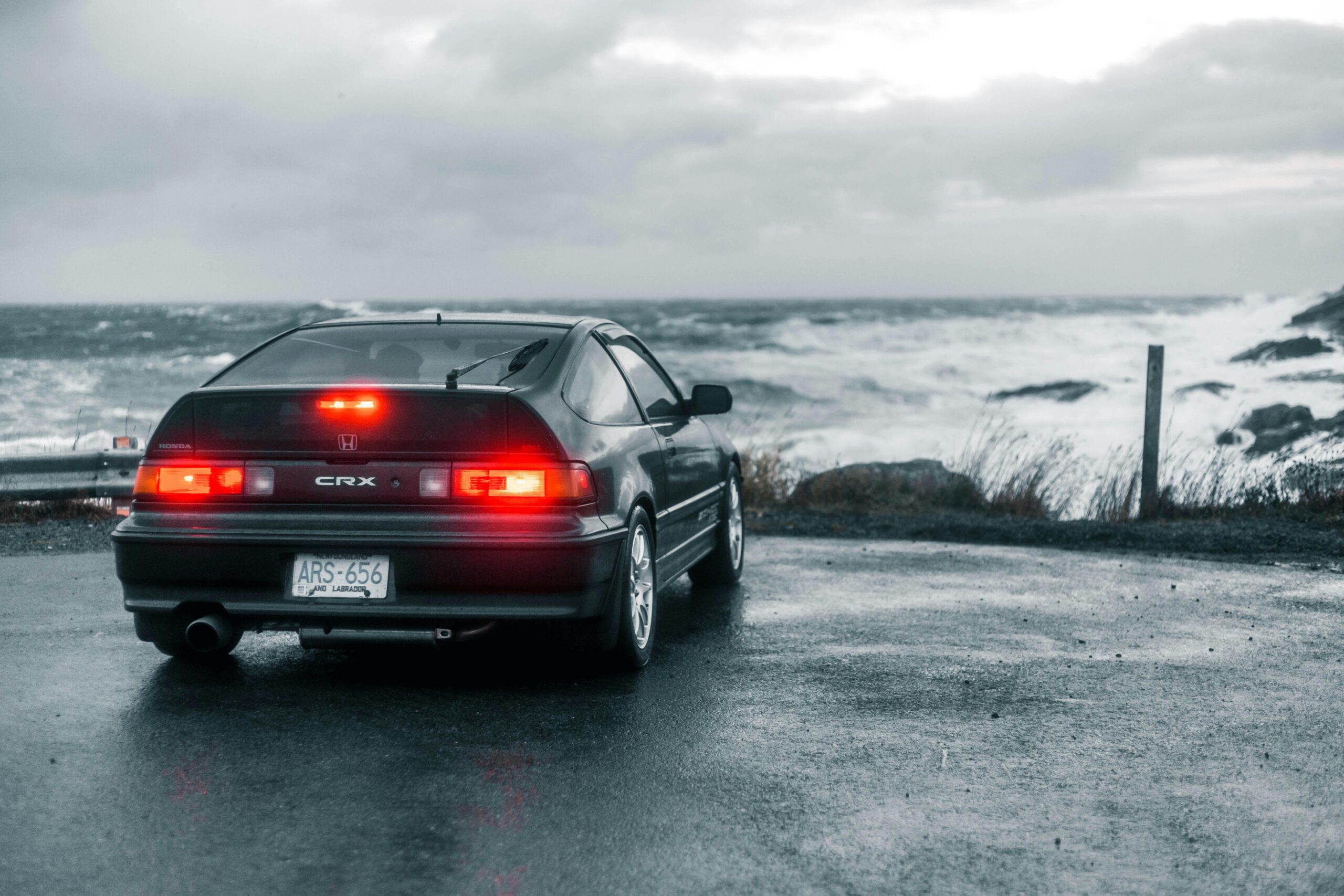 A Honda CRX parked by a stormy ocean, highlighting its taillight against the gloomy seascape.