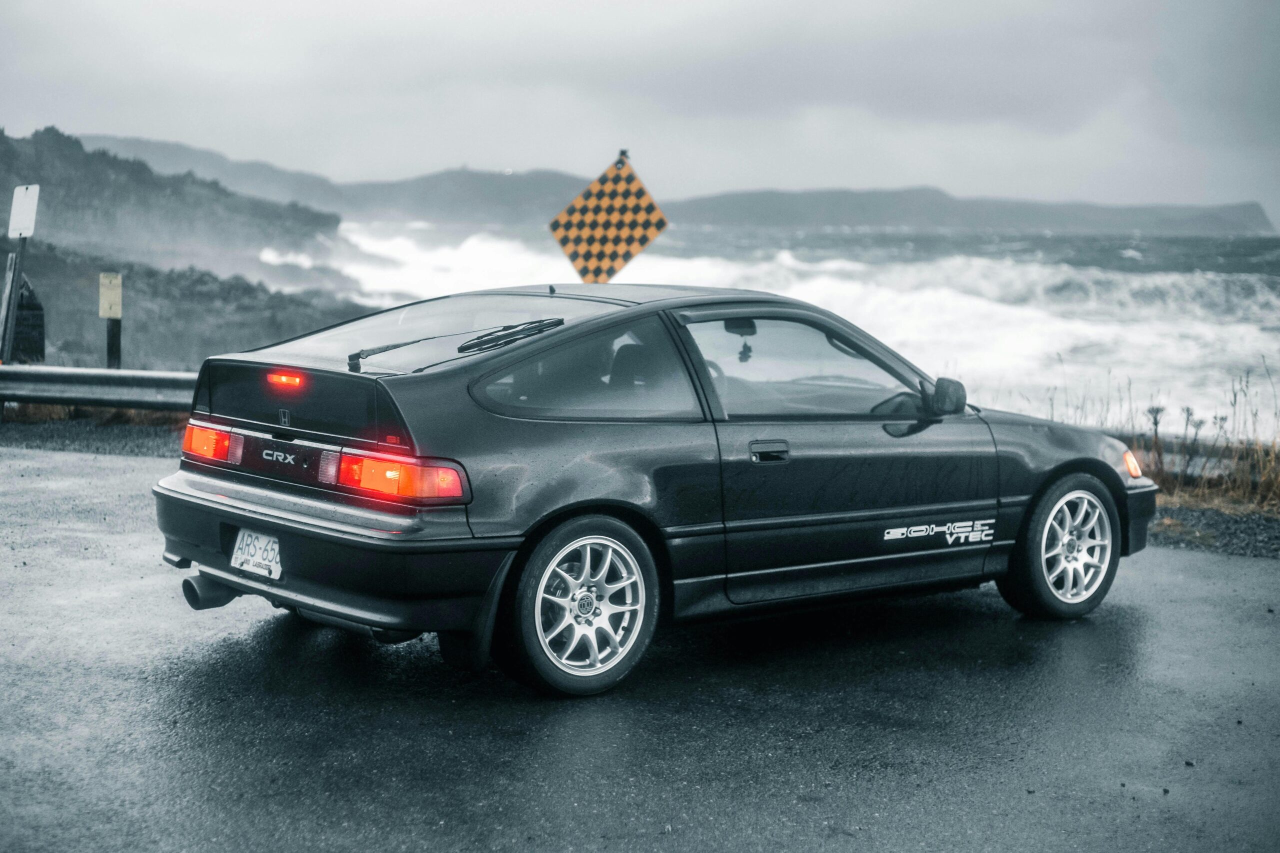 A Honda CRX parked by the coastline on a rainy day with rough sea and waves in the background.